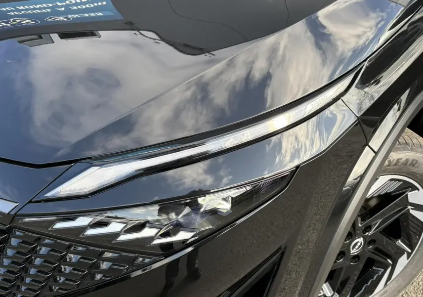 Gros plan sur le phare avant gauche et la jante noire du Nissan Qashqai III e-Power 2025 en peinture métallisée noire.