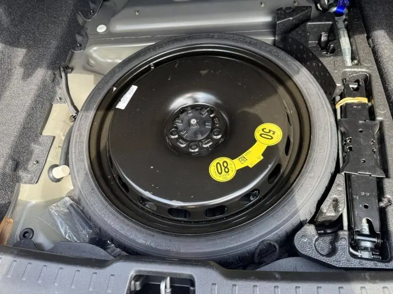 Vue rapprochée de la roue de secours noire avec étiquette jaune dans le coffre d’un Volvo S60 gris Brume Métallisé 2024.