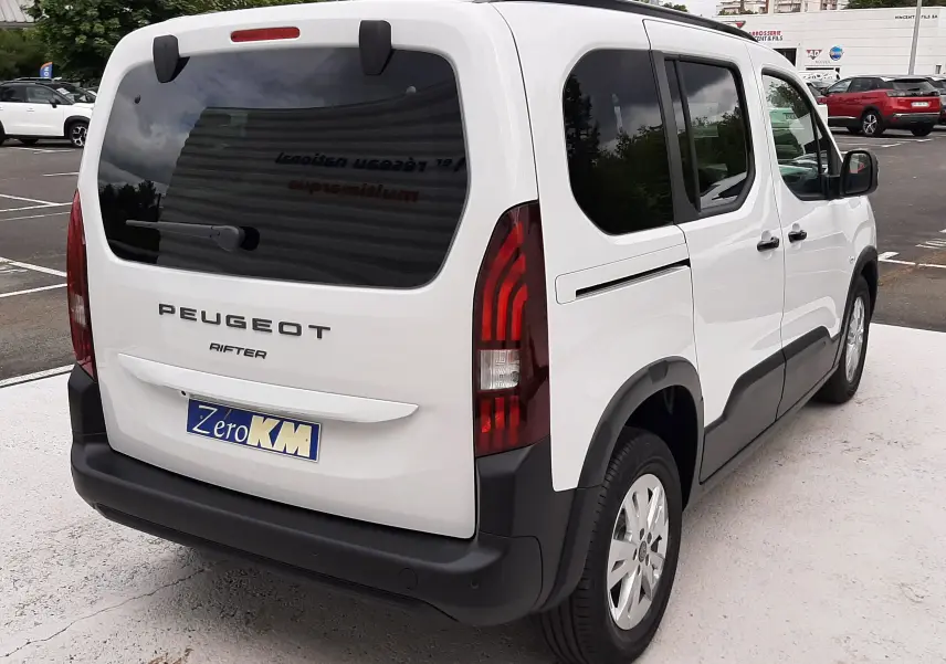 Vue 3/4 arrière droite d'un Peugeot Rifter blanc Kaolin avec vitres teintées et protections noires sur un parking.