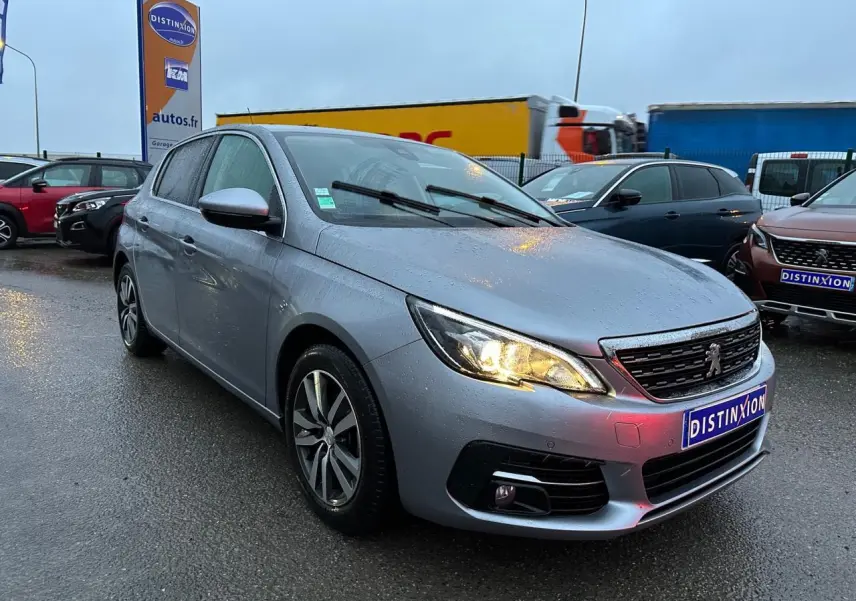 PEUGEOT 308 gris Artense en 3/4 avant droit sous la pluie, phare allumé et jantes alliage visibles.