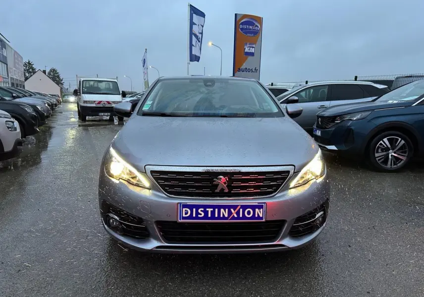 Peugeot 308 gris Artense vue de face, phares allumés, sur parking humide avec panneau Distinxion en arrière-plan.