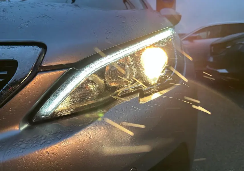 Gros plan sur le phare avant droit allumé d’une Peugeot 308 gris Artense, avec gouttes d’eau sur la carrosserie.