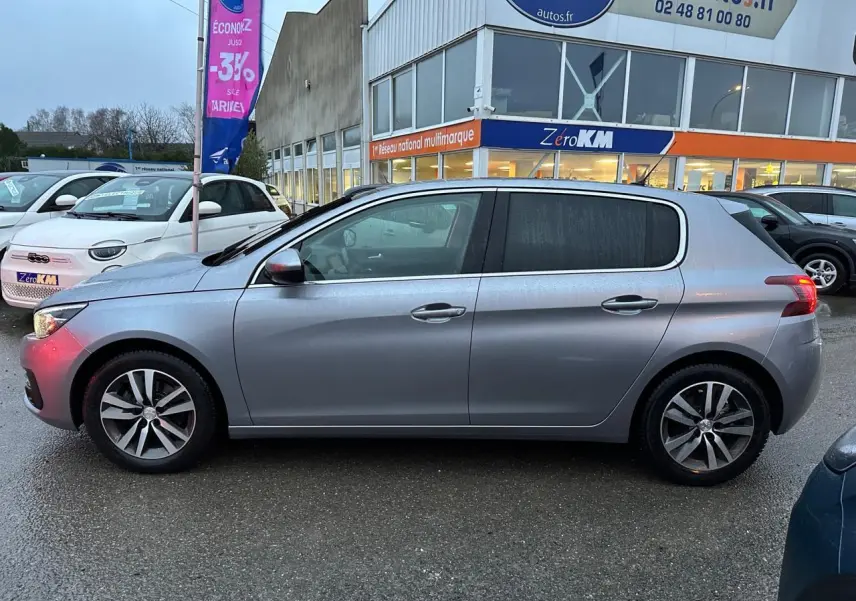 Peugeot 308 gris Artense vue de profil côté gauche, avec jantes alliage et feux arrière allumés.