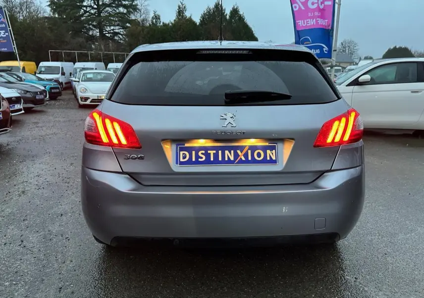 Vue arrière d'une Peugeot 308 gris Artense avec feux arrière allumés sur un parking humide.