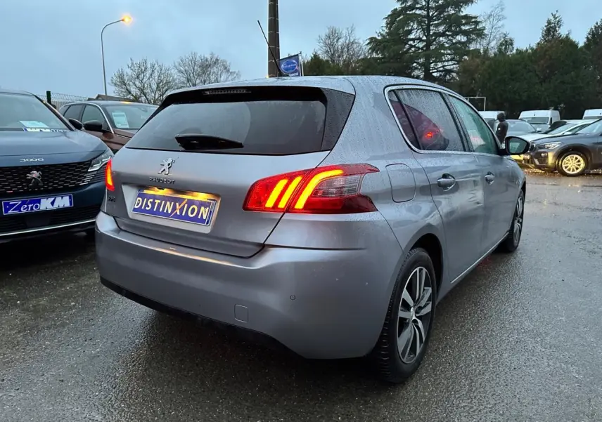 Peugeot 308 gris Artense vue 3/4 arrière droit avec feux arrière allumés sur parking humide.