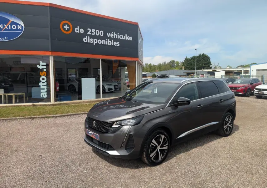 Peugeot 5008 gris métallisé vue 3/4 avant droit avec jantes bi-ton et calandre GT devant un concessionnaire.
