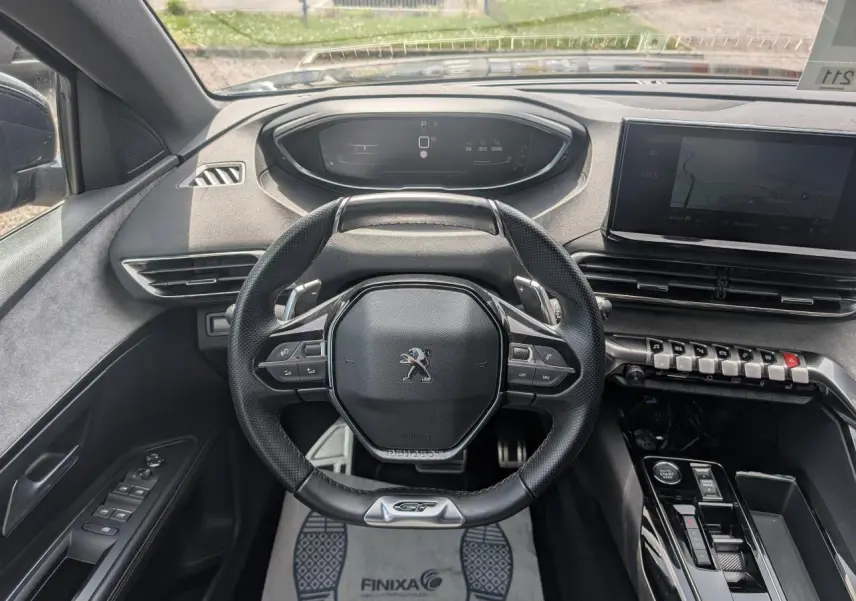 Vue intérieure frontale du volant cuir perforé noir avec badge GT du Peugeot 5008 gris 2023, tableau de bord et écran tactile visibles.