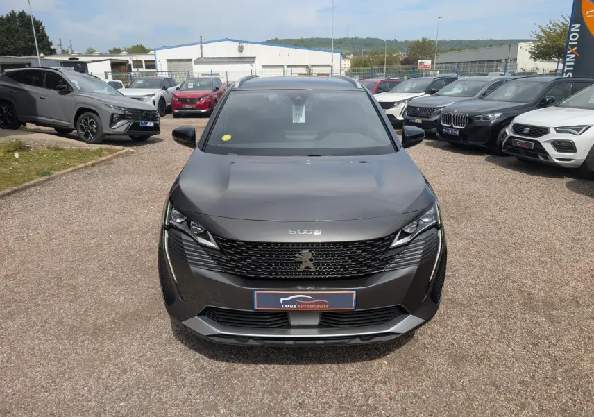 Peugeot 5008 gris métallisé vue de face, avec calandre GT noire et feux LED 3D, sur parking extérieur.
