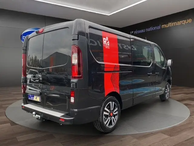 Vue 3/4 arrière droite du Renault Trafic noir avec jantes alu noires, barres de toit et attelage visible en intérieur showroom.