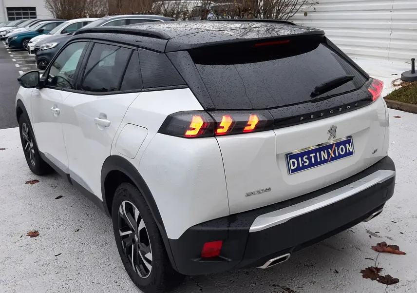 Vue 3/4 arrière droite d'un Peugeot 2008 blanc nacré avec toit noir et feux arrière allumés sur parking extérieur.