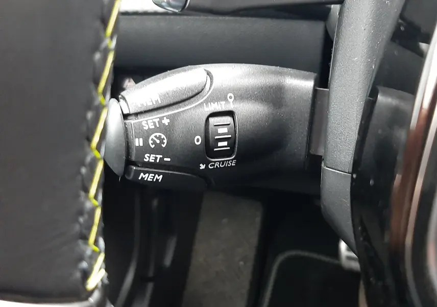 Gros plan sur la commande de régulateur de vitesse au volant cuir noir avec surpiqûres jaunes d'une Peugeot 2008 blanche nacrée 2023.