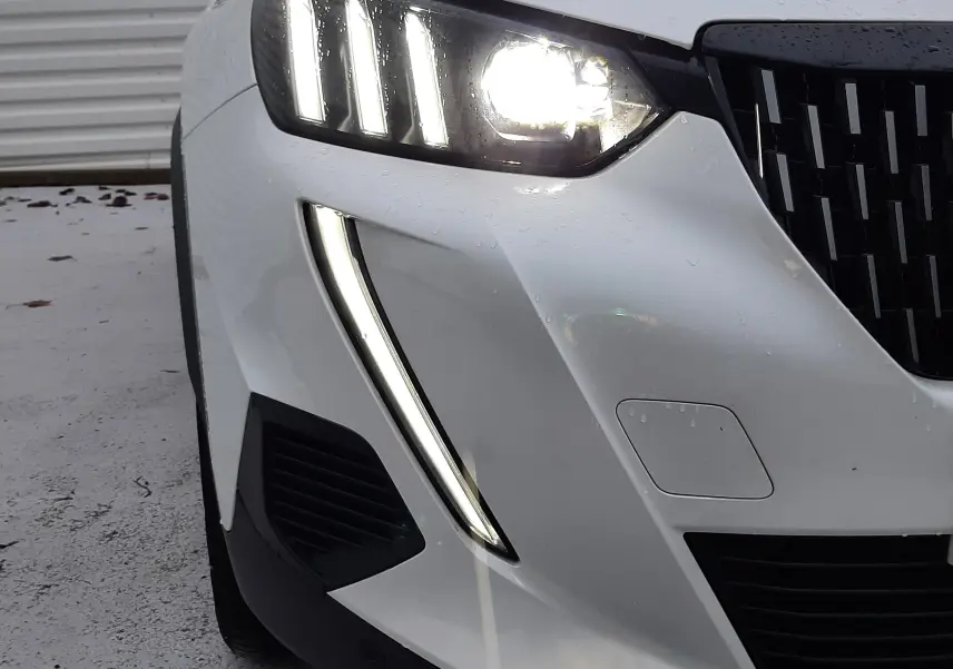 Gros plan sur l'avant droit du Peugeot 2008 blanc nacré avec phares LED allumés et calandre noire.