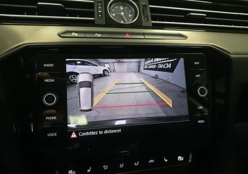 Écran tactile de la Volkswagen Passat SW gris montrant la caméra de recul avec lignes de guidage colorées et horloge analogique au-dessus.