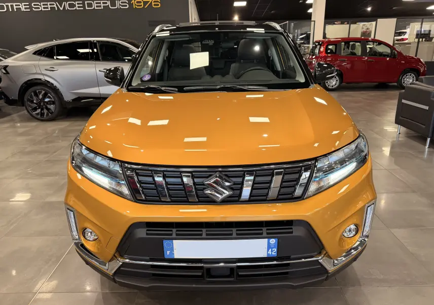Vue avant d'un Suzuki Vitara 1.5 Dualjet Hybrid jaune avec toit noir, en showroom lumineux, calandre chromée visible.