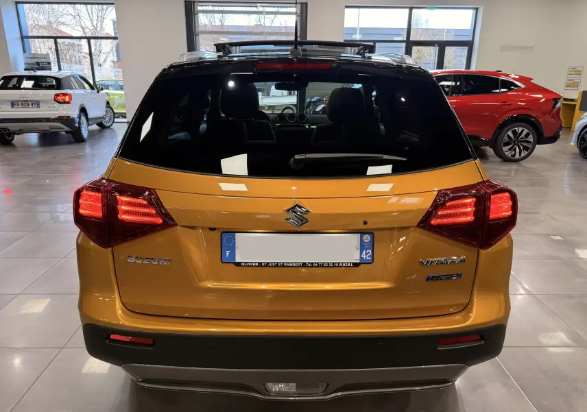 Vue arrière d'un Suzuki Vitara 1.5 Dualjet Hybrid jaune avec toit noir, feux arrière allumés en concession.