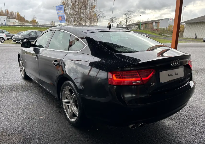 Audi A5 Sportback noir vue 3/4 arrière droit sous la pluie avec feux arrière allumés et jantes alliage.