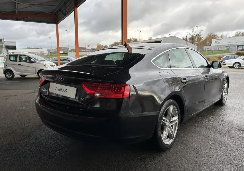 Audi A5 Sportback noir vue 3/4 arrière droit sous abri, avec feux arrière allumés et jantes alliage cinq branches.