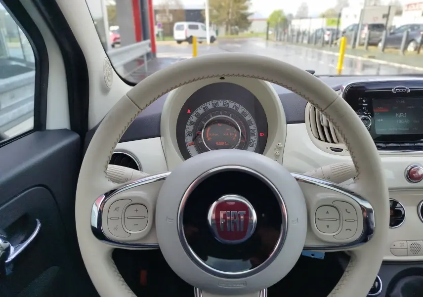 Vue intérieure centrée sur le volant beige de la Fiat 500 blanche avec tableau de bord et écran multimédia visibles.