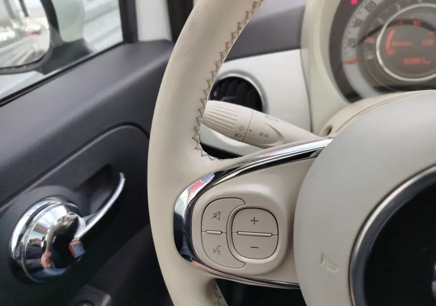 Gros plan sur le volant beige et la commande audio d’une FIAT 500 Bossa Nova White Pastel, tableau de bord partiellement visible.