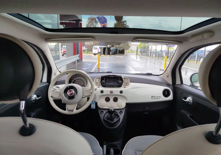 Vue intérieure avant de la Fiat 500 blanche avec tableau de bord épuré, volant beige et toit panoramique ouvert.