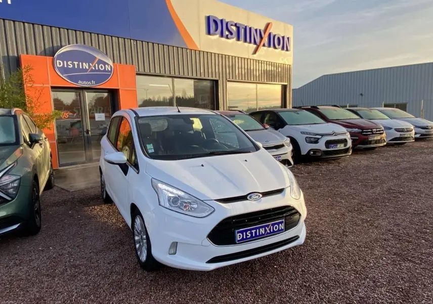 Ford B-MAX blanc vue 3/4 avant droit sur un parking devant un concessionnaire Distinxion au coucher du soleil.