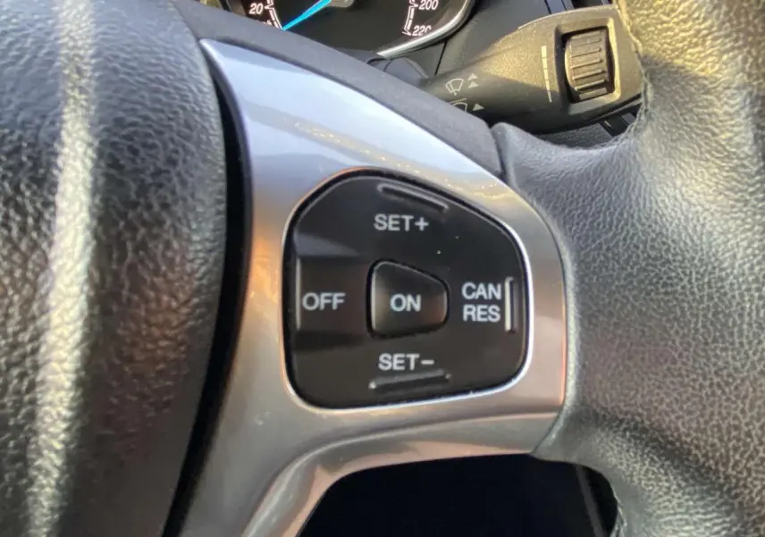 Gros plan sur les commandes du régulateur de vitesse au volant d'une Ford B-MAX blanche, tableau de bord en arrière-plan.