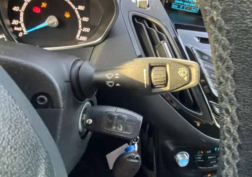Gros plan sur la colonne de direction et le tableau de bord d’une Ford B-MAX blanche de 2014, avec clé insérée et commandes d’essuie-glace visibles.