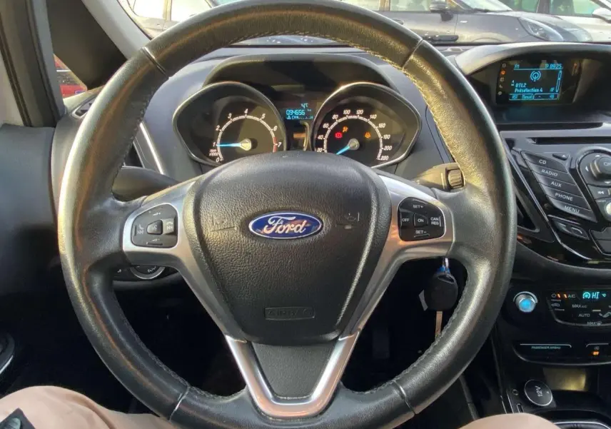 Vue intérieure centrée sur le volant cuir noir de la Ford B-MAX blanche, avec tableau de bord et commandes visibles.