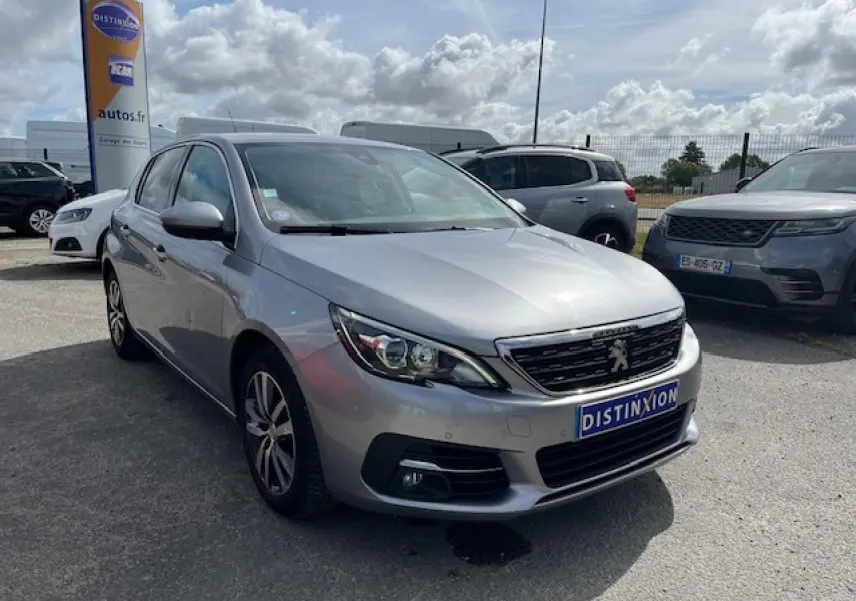 Vue 3/4 avant d'une Peugeot 308 gris clair métallisé garée sur un parking extérieur sous ciel partiellement nuageux.