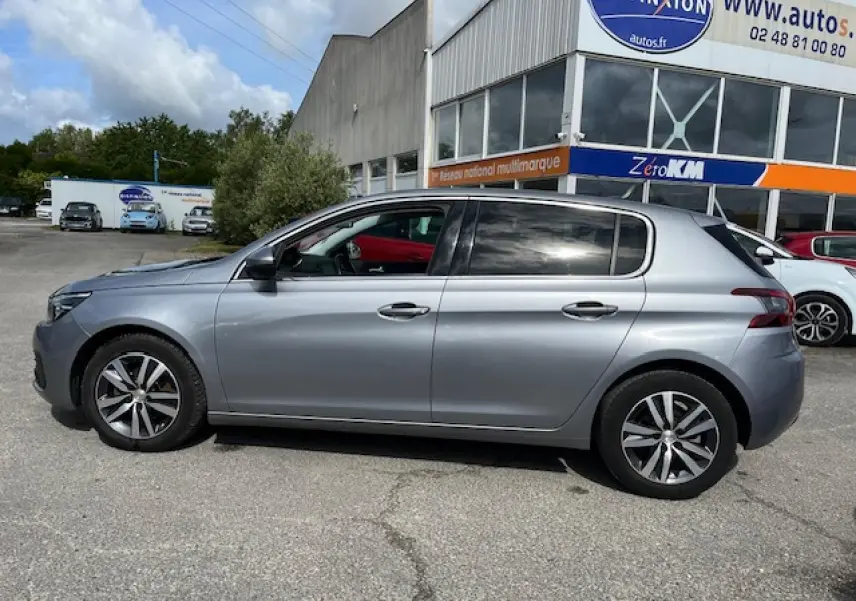 Profil droit d'une Peugeot 308 gris clair métallisé garée devant un bâtiment commercial, avec jantes alliage visibles.