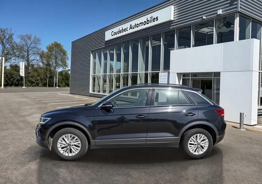 Profil côté gauche d’un Volkswagen T-Roc noir 2025 garé devant un bâtiment moderne de concession automobile.