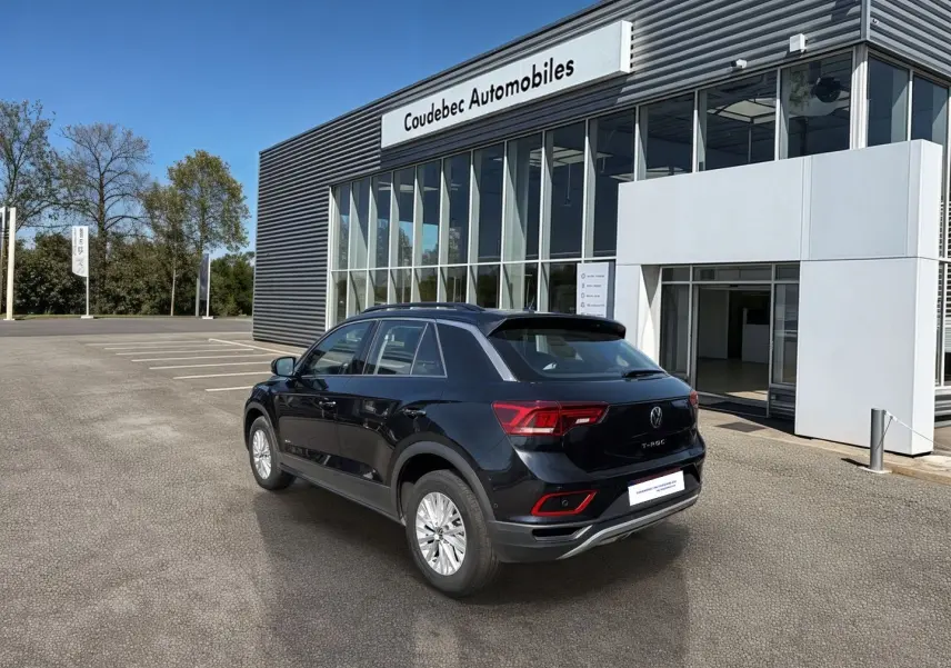 Volkswagen T-Roc noir vue 3/4 arrière droit, stationné devant un bâtiment moderne de concession automobile.