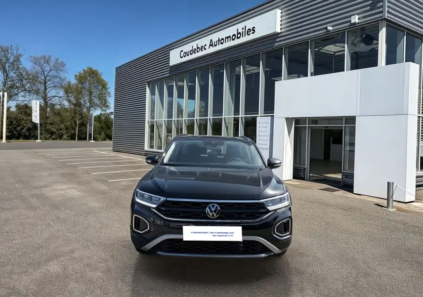 Vue frontale d'un Volkswagen T-Roc noir 2025 garé devant un bâtiment moderne de concession automobile.