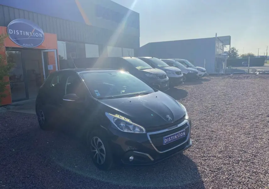 Peugeot 208 noire vue 3/4 avant droit sur parking gravillonné devant un bâtiment commercial.