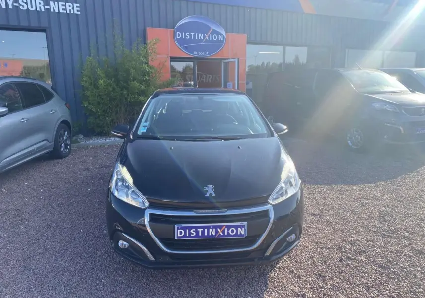 Peugeot 208 noire vue de face, avec calandre chromée et phares allumés, stationnée devant un garage.
