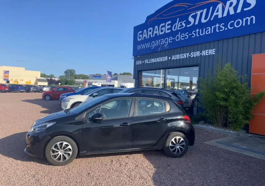 Peugeot 208 noire vue de profil côté gauche, avec jantes acier 15 pouces et roue de secours, stationnée devant un garage.