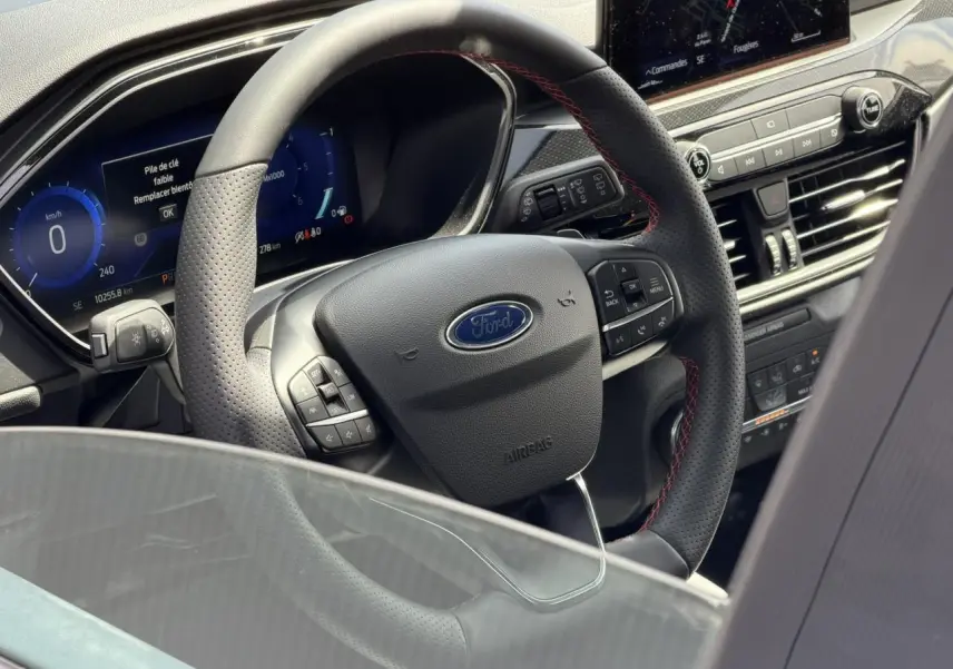 Vue intérieure centrée sur le volant noir perforé avec surpiqûres rouges du Ford Kuga 2023 gris Solar, tableau de bord numérique visible.