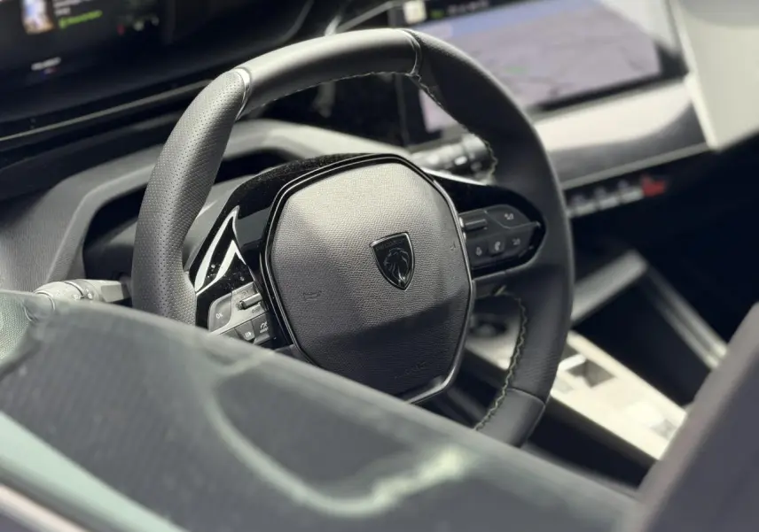 Gros plan sur le volant compact cuir noir avec logo Peugeot, tableau de bord numérique et écran tactile en arrière-plan.
