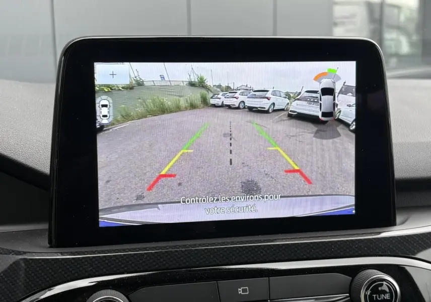 Écran de caméra de recul montrant l’arrière d’un Ford Kuga gris graphite en stationnement, avec lignes de guidage colorées.