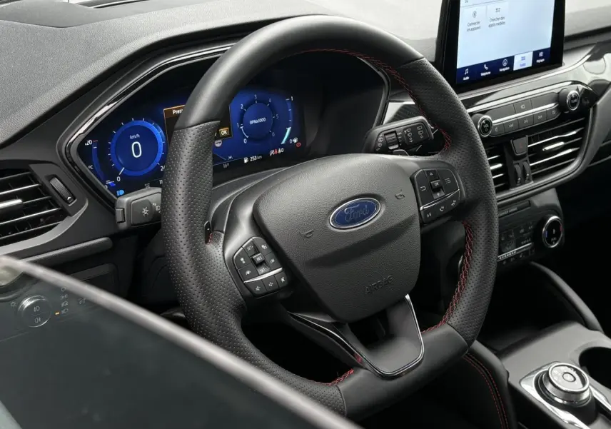 Vue intérieure centrée sur le volant noir perforé avec surpiqûres rouges du Ford Kuga 2023 et tableau de bord numérique bleu.