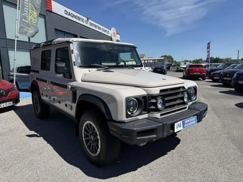 Vue 3/4 avant droit d'un INEOS Grenadier beige 2024 avec calandre noire et phares ronds, stationné en extérieur.