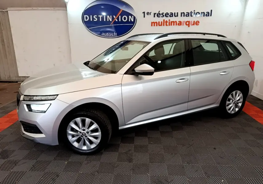 Vue de profil côté gauche d'un Skoda Kamiq gris clair 2022 en showroom avec jantes alliage et barres de toit noires.