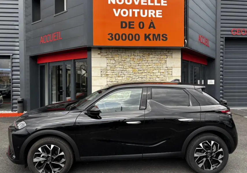 Vue profil côté gauche d'une DS3 noire 2024 avec jantes alliage et toit noir, stationnée devant un bâtiment commercial.