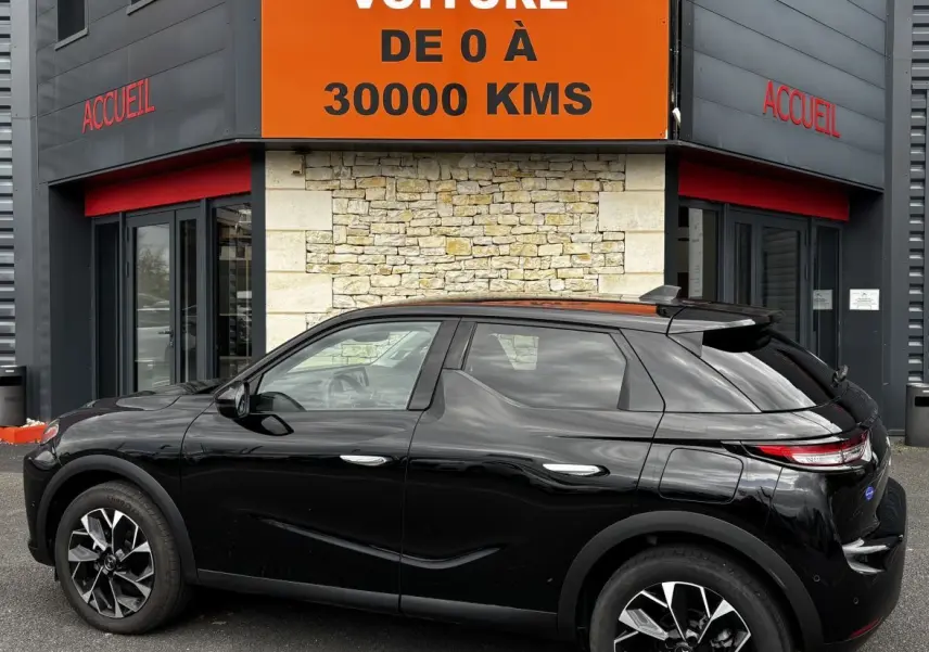 Vue latérale droite d'une DS3 noire 2024 garée devant un bâtiment avec un grand panneau orange "Votre nouvelle voiture de 0 à 30000 kms".