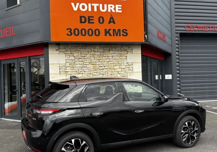 Vue 3/4 arrière droite d'une DS3 noire 2024 avec jantes alliage noires et toit noir devant un bâtiment commercial.
