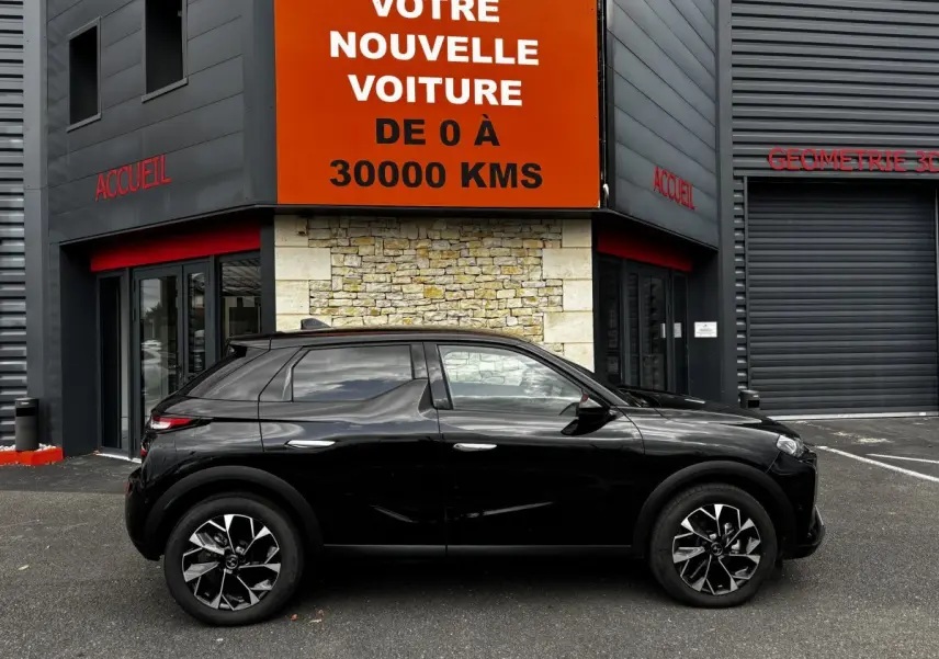 DS3 noire vue de profil côté gauche, avec jantes noires et détails chromés, devant un bâtiment commercial.