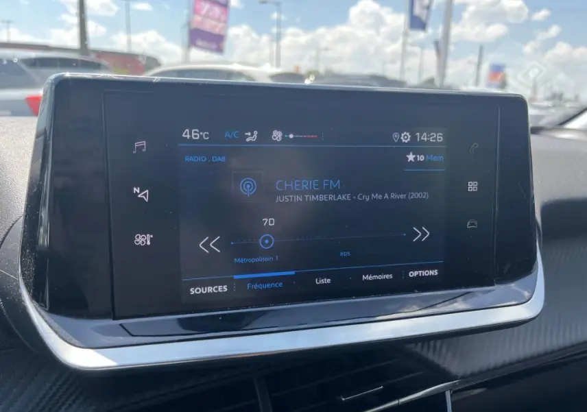 Écran tactile central de la Peugeot 208 gris clair 2023, affichant la radio et les commandes climatisation.