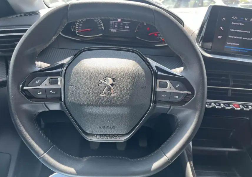 Vue rapprochée du volant noir de la Peugeot 208 2023, avec tableau de bord et écran tactile visibles en arrière-plan.