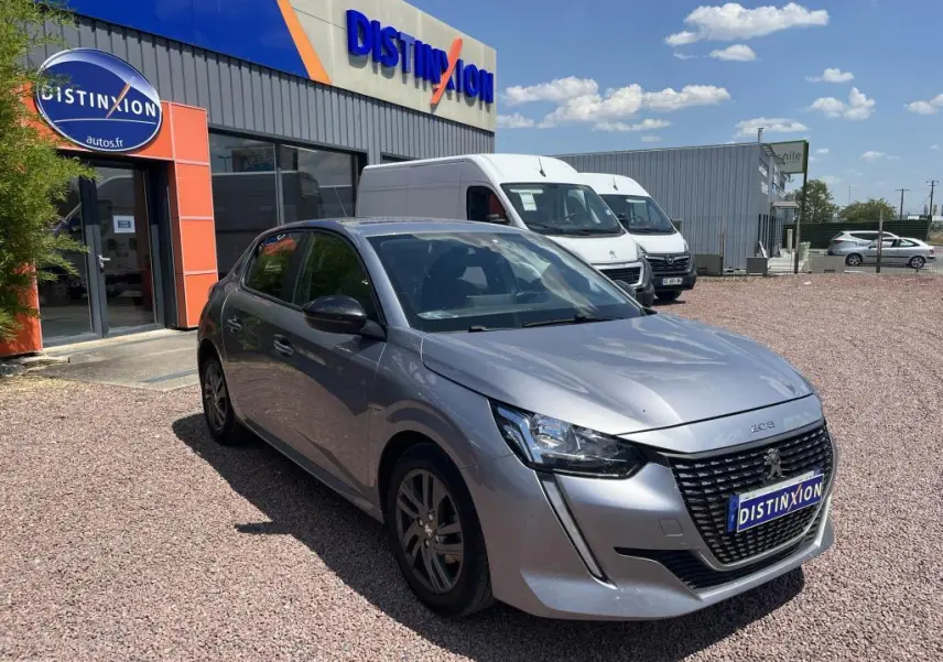 Peugeot 208 gris clair vue 3/4 avant droit, avec calandre noire distinctive, stationnée devant un garage Distinxion.