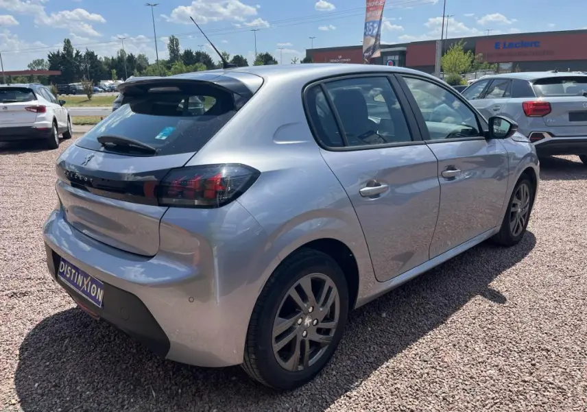 Peugeot 208 gris clair vue 3/4 arrière droit, avec feux arrière noirs et jantes alliage, stationnée sur gravier.